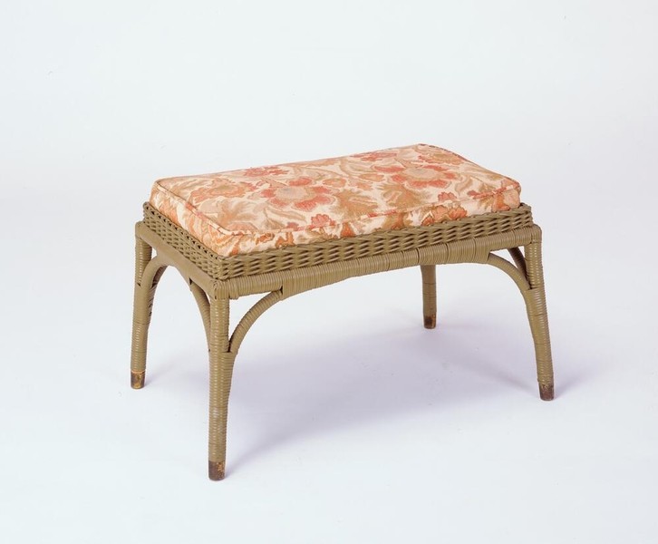 Greene and Greene designed footstool from Aunt Julia's bedroom at the Gamble House, featuring a natural wicker construction with woven seat platform, curved legs with decorative cutouts, cross-stretchers, and an upholstered cushion in cream fabric with stylized floral pattern in coral-pink and golden tones, demonstrating the brothers' use of lighter wicker furniture for bedroom spaces to create a more airy and feminine aesthetic