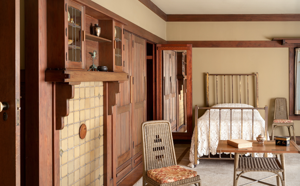 Aunt Julia Huggins' bedroom at the Gamble House showing metal bed with vertical slat headboard and footboard and white crocheted bedspread at left, natural wicker chaise lounge with high rounded back and floral upholstered cushion in coral-pink tones at right, natural wicker side chair with geometric cutout pattern and floral cushion at center, Honduras mahogany occasional table with fresh orchids in ceramic bowl and framed photograph, dressing table with tilting mirror visible in background, windows with sheer curtains, warm golden-yellow walls with Honduras mahogany trim, exposed beam ceiling with dark-stained beams, Arts and Crafts wall sconces with amber glass shades, and cream-colored carpet, creating a light and airy feminine space distinct from the darker wood tones elsewhere in the house