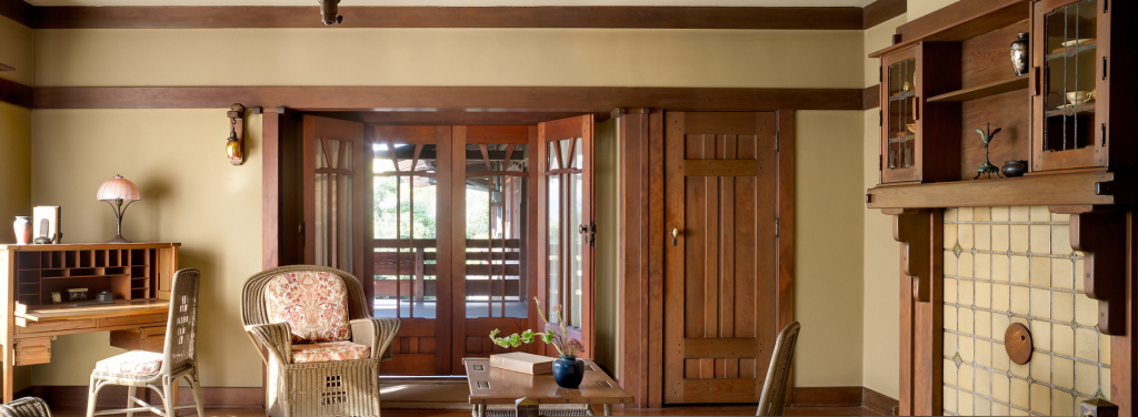 Alternate view of Aunt Julia Huggins' bedroom at the Gamble House showing the Honduras mahogany doors to the sleeping porch with V-shaped muntins in the upper glass panels and sheer curtains, soft green painted wicker furniture including easy chair with floral upholstered cushions and footstool at left, writing desk with tilting mirror and pink table lamp at far left, built-in fireplace with cream-colored tile surround and Honduras mahogany cabinetry at right, exposed beam ceiling with dark-stained beams and cream plaster between, cream-colored walls with mahogany trim, and natural light streaming through the porch doors, creating a light and airy feminine space