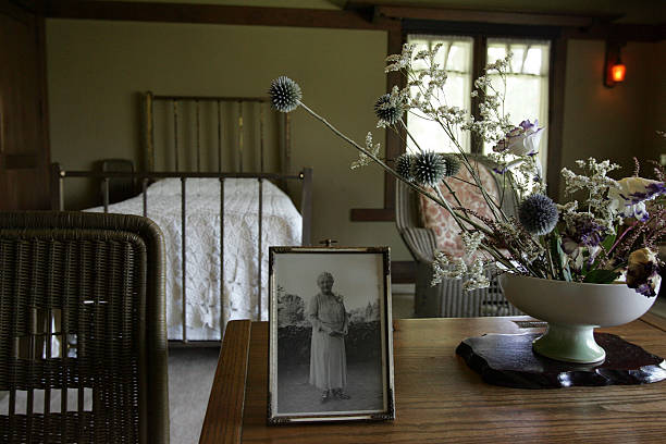 Detail view of Aunt Julia Huggins' bedroom at the Gamble House showing a Honduras mahogany occasional table with fresh flowers in a white ceramic vase and a framed vintage photograph of an elderly woman in white dress standing outdoors, natural wicker side chair with geometric cutout pattern in the back at left, metal bed with vertical slat headboard and white coverlet visible in the background, windows with V-shaped muntins and sheer curtains providing soft natural light, and warm cream-colored walls, creating an intimate personal space with period furnishings and family mementos