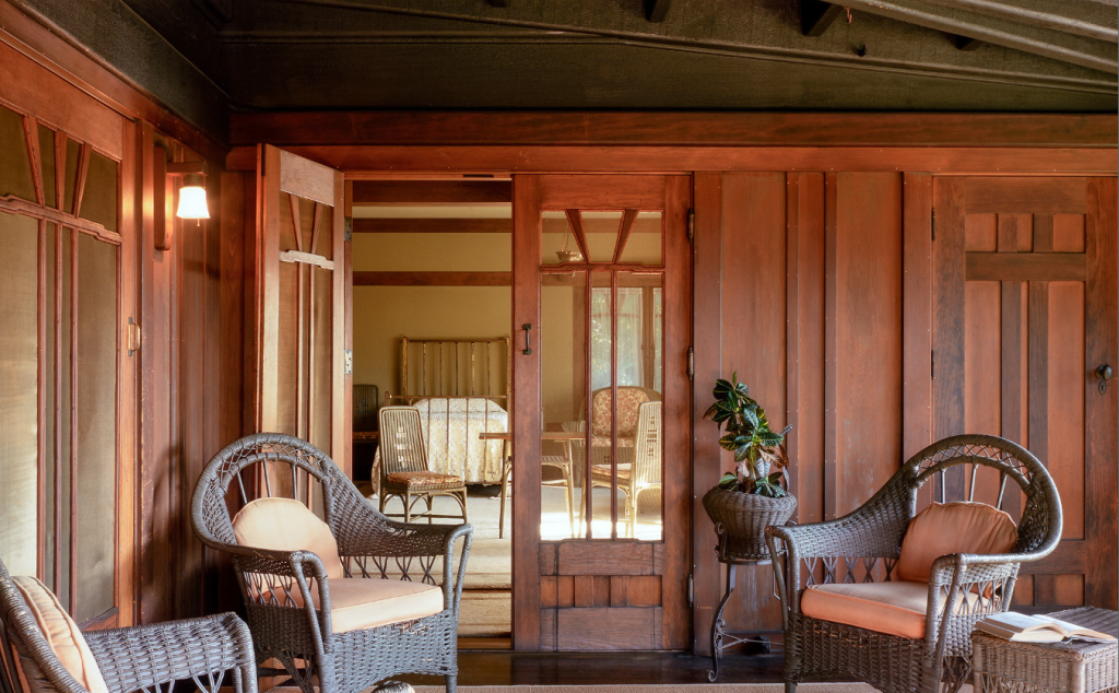 Aunt Julia Huggins' sleeping porch at the Gamble House showing the Honduras mahogany door with V-shaped muntins in the upper glass panel opening to the bedroom where the metal bed with vertical slat headboard and white crocheted bedspread is visible, natural wicker furniture on the sleeping porch including two high-backed chairs with rounded fan-shaped backs and coral-pink upholstered cushions flanking the doorway, wicker side chair visible through the doorway, potted plant in wicker basket at right, Honduras mahogany wall paneling with vertical board-and-batten design, exposed beam ceiling painted soft green, Arts and Crafts wall sconce with amber glass shade at left, and warm golden afternoon light streaming through the space, demonstrating the indoor-outdoor living philosophy central to the California bungalow lifestyle