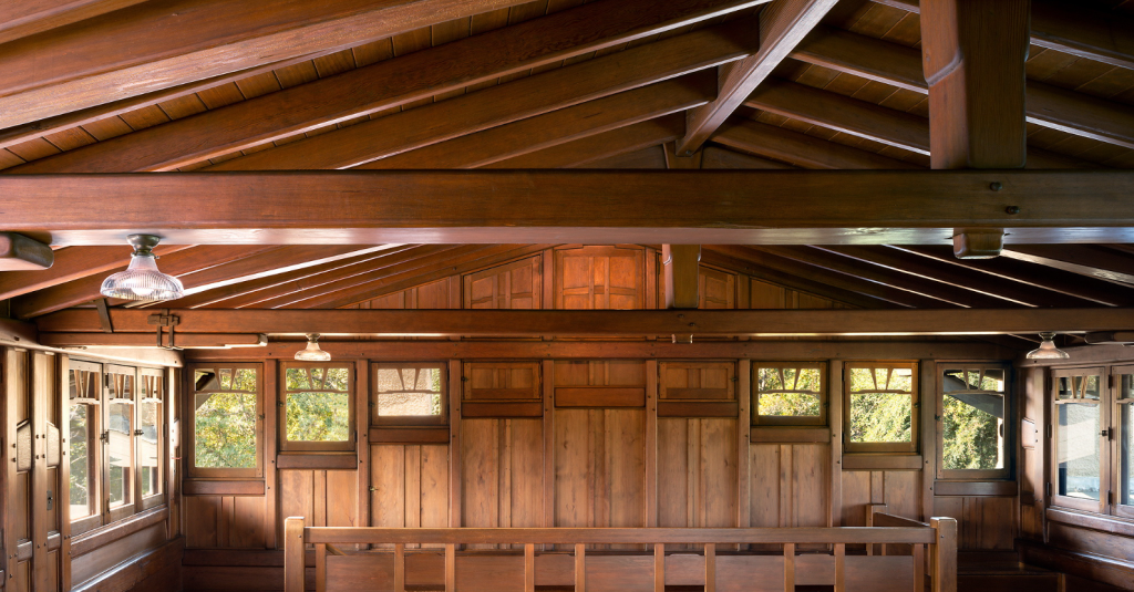 Panoramic view of the Gamble House attic space showing the dramatic exposed structural system with massive Honduras mahogany rafters, beams, and purlins creating a complex geometric pattern against the tongue-and-groove wood ceiling, natural light streaming through casement windows with V-shaped muntins on both sides, built-in storage cabinets with geometric panel designs below the windows, vertical wood paneling on the walls, built-in teak bench with horizontal slat back at center, period light fixtures with glass shades mounted on the ceiling and walls, hardwood flooring, and the warm golden-brown tones throughout, demonstrating how Greene and Greene transformed a utilitarian attic into an architecturally significant space through meticulous craftsmanship and exposed structural expressiveness