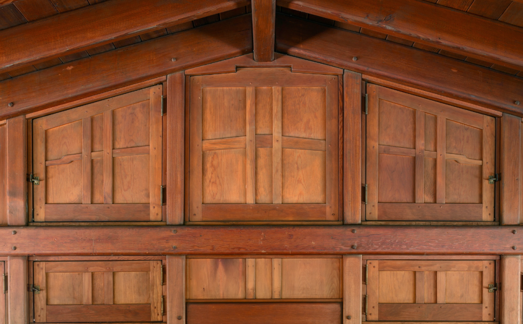 Close-up detail view of built-in storage cabinets in the Gamble House attic showing two tiers of custom Honduras mahogany cabinet doors with geometric panel designs featuring vertical and horizontal divisions, brass hinges and latches, exposed rafters and tongue-and-groove ceiling above, massive horizontal beams with square pegs, and the warm golden-brown patina of aged wood throughout, demonstrating Greene and Greene's meticulous attention to storage design even in utilitarian attic spaces