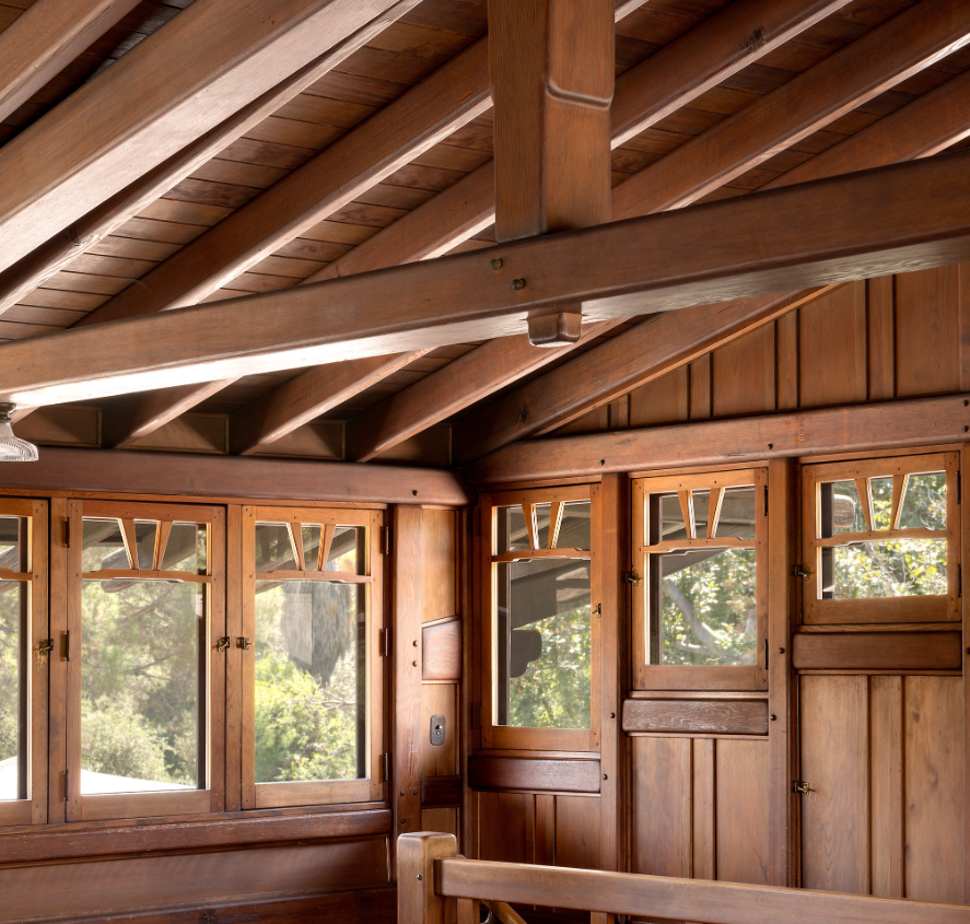 Interior view of the attic space at the Gamble House photographed by Alexander Vertikoff, showing the dramatic exposed structural system with massive Honduras mahogany rafters, beams, and purlins creating a complex geometric pattern against the tongue-and-groove wood ceiling, natural light streaming through casement windows with V-shaped muntins on both sides, built-in storage cabinets with geometric panel designs flanking the windows, vertical wood paneling on the walls, hardwood flooring, and the warm golden-brown tones of the aged wood throughout, demonstrating Greene and Greene's philosophy of making even utilitarian spaces architecturally significant through exposed structural expressiveness and meticulous craftsmanship