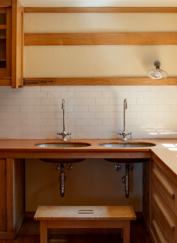 Butler's pantry at the Gamble House showing dual copper sinks with hammered finish set into a Honduras mahogany counter, nickel-plated faucets with cross handles mounted on the white subway tile backsplash laid in running bond pattern, open shelving above with Honduras mahogany supports and cream-colored plaster walls between the horizontal bands of wood trim, industrial-style pendant light fixture with white porcelain shade and exposed metal hardware mounted on the right, Honduras mahogany cabinetry below the counter with geometric panel designs, exposed plumbing with chrome traps visible beneath the sinks, and a small wooden step stool positioned under the counter for reaching upper shelves, demonstrating the functional efficiency and honest expression of materials characteristic of Greene and Greene's service spaces where form follows function while maintaining the aesthetic integrity found throughout the house