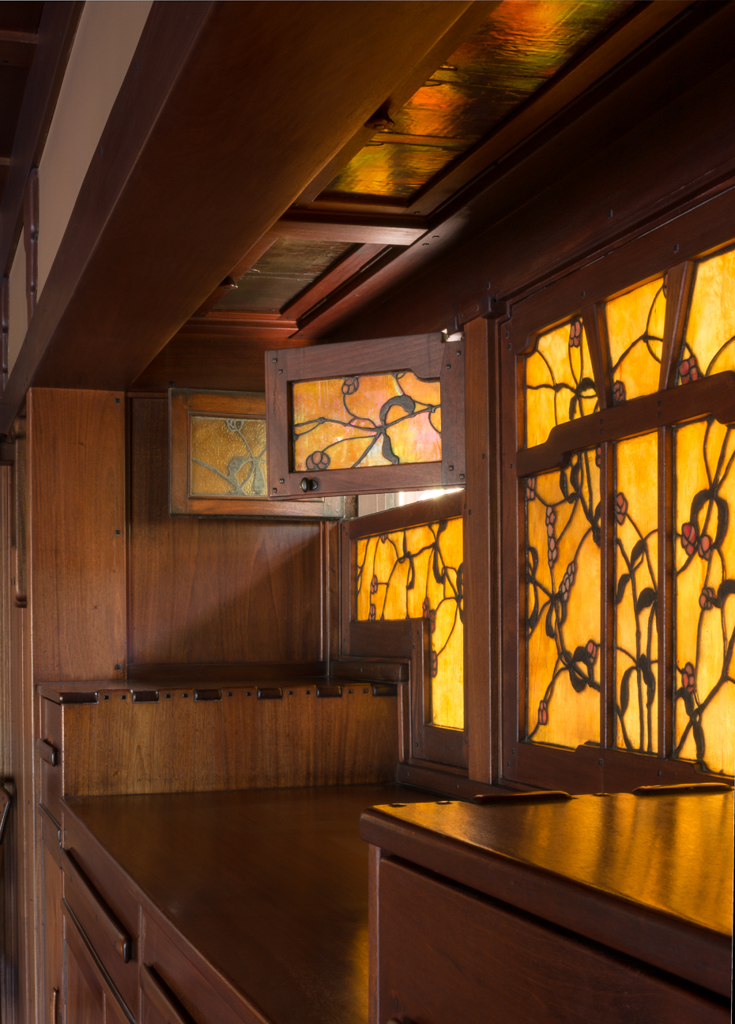 Detail view of the dining room built-in sideboard at the Gamble House showing the backlit Tiffany Studios stained glass panels with stylized oak tree branches featuring gnarled trunks and organic forms in golden-yellow and amber glass with darker leading creating silhouettes, Honduras mahogany cabinetry with geometric panel designs, small decorative glass panel with bird motif at upper left, open shelving displaying pottery, cloud-lift copper drawer pulls, and the warm glow of light filtering through the art glass creating patterns on the mahogany surfaces, demonstrating the integration of decorative arts with functional cabinetry