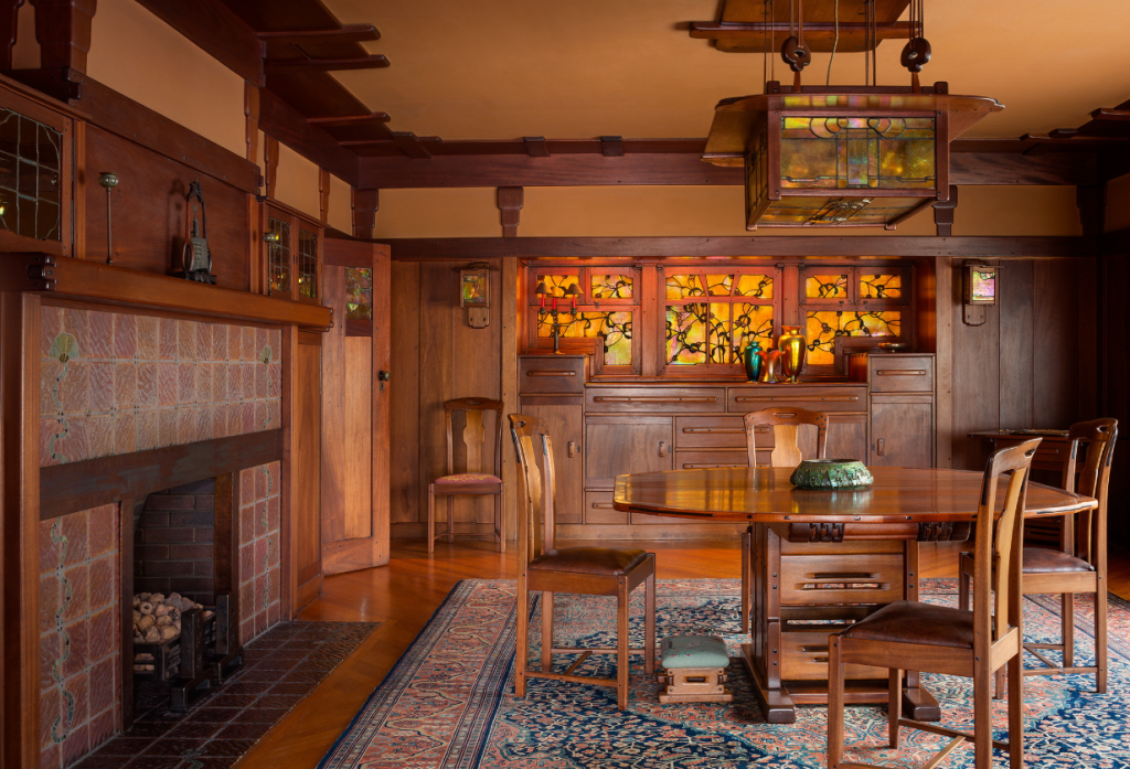 Dining room of the Gamble House showing the built-in Honduras mahogany sideboard with backlit Tiffany Studios stained glass panels depicting stylized oak tree branches in golden-yellow and amber glass, multiple drawers and cabinet doors with cloud-lift copper pulls, Arts and Crafts wall sconces flanking the sideboard, custom dining table by Peter Hall Manufacturing Company with bird's-eye maple inlay and ebony pegs surrounded by dining chairs with cloud-lift cutout handles and upholstered seats, built-in fireplace with iridescent tile surround and Honduras mahogany mantel at left, exposed beam ceiling with dark-stained beams and cream plaster between creating a coffered effect, custom pendant light fixture with geometric design suspended from the ceiling, Arts and Crafts carpet with floral and geometric patterns, and warm golden lighting creating an intimate dining atmosphere