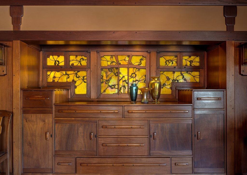 Front view of the built-in dining room sideboard at the Gamble House crafted by Peter Hall Manufacturing Company in Honduras mahogany, featuring backlit Tiffany Studios stained glass panels in the upper section depicting stylized oak tree branches with gnarled trunks in golden-yellow and amber glass arranged in a symmetrical composition across multiple panels, multiple drawers with cloud-lift copper pulls and decorative escutcheons, cabinet doors with figured wood panels and copper hardware, flanked by additional storage cabinets on both sides, massive Honduras mahogany beam above supported by cloud-lift brackets, iridescent pottery vases in green, amber, and golden tones displayed on the counter, and the warm glow of backlighting illuminating the art glass creating a focal point for the dining room