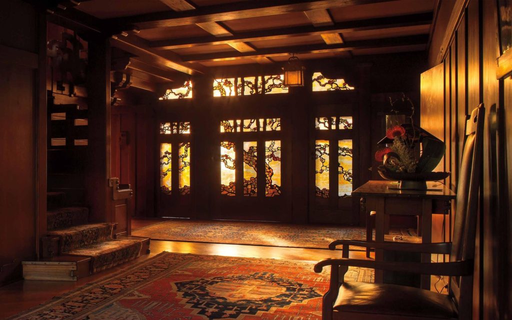 Entrance hall of the Gamble House at sunrise photographed by Alexander Vertikoff showing the dramatic Tiffany Studios stained glass entry doors with stylized oak tree branches in golden-yellow, amber, and green glass glowing with warm morning light, Honduras mahogany wall paneling, exposed beam ceiling with dark-stained beams and cream plaster between, built-in bench with upholstered cushions in floral pattern at left, grand staircase with cloud-lift balustrade visible at left, Arts and Crafts carpet with geometric and floral patterns in reds, blues, and golds, Honduras mahogany hall table at right, and the warm golden glow throughout creating a welcoming and contemplative entrance space