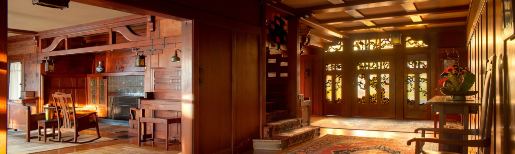 Panoramic view of the Gamble House entrance hall at sunrise showing the complete spatial composition with Tiffany Studios stained glass entry doors glowing with golden morning light depicting stylized oak tree branches, Honduras mahogany wall paneling throughout, exposed beam ceiling with dark-stained beams creating a coffered pattern, built-in bench with upholstered cushions at right, grand staircase with cloud-lift balustrade visible at left, views through doorways into the dining room at left showing the fireplace and built-in sideboard with backlit Tiffany glass, Arts and Crafts carpets with floral and geometric patterns, hardwood flooring, and the warm golden glow of sunrise streaming through the art glass creating a welcoming and contemplative entrance that connects the major living spaces of the house
