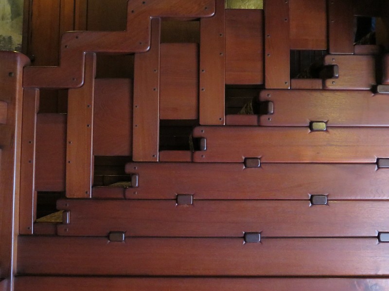 Close-up detail of the grand staircase handrail at the Gamble House showing the cloud-lift balustrade design with stepped geometric forms descending in a rhythmic pattern, Honduras mahogany construction with square ebony pegs arranged in horizontal rows across each panel, the sculptural quality of the organic curved forms creating a visual rhythm that echoes the ascending stairs, and the meticulous joinery demonstrating Greene and Greene's philosophy of making structural connections visible and decorative