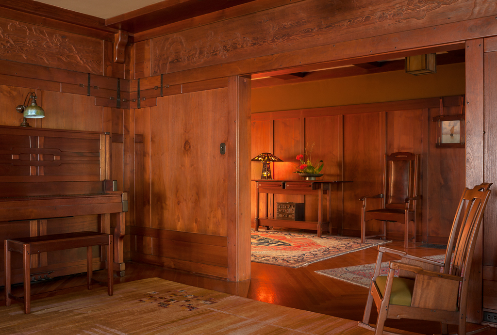 View from the entrance hall into the living room of the Gamble House showing Honduras mahogany wall paneling with vertical board-and-batten design, custom Honduras mahogany pocket doors with geometric panel designs, built-in serving table or console at left with lower shelf, Arts and Crafts wall sconce with amber glass shade, hardwood flooring, Arts and Crafts carpet with floral and geometric patterns, view into the living room showing table lamp with amber glass shade, fresh flowers in ceramic vase, Greene and Greene furniture including chairs with vertical slats, and warm golden lighting creating intimate connected spaces that flow together while maintaining distinct functions