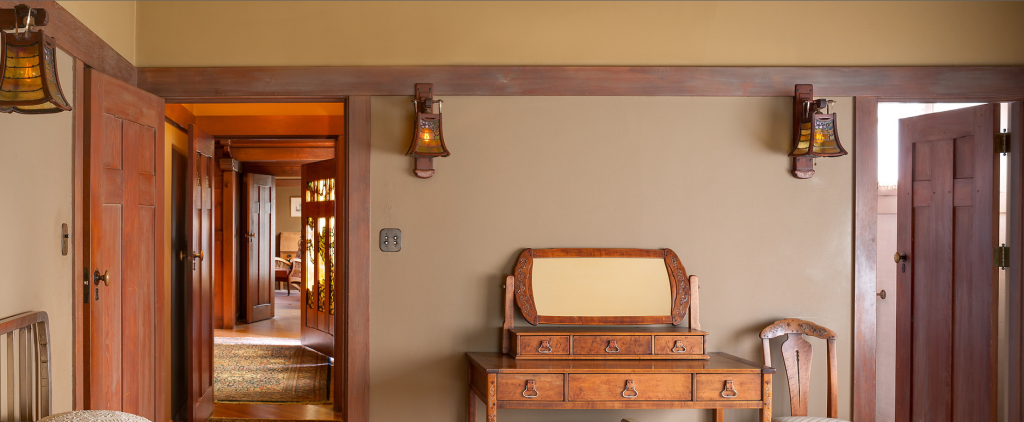 Guest bedroom at the Gamble House showing view through Honduras mahogany pocket doors with geometric panel designs into the hallway beyond where Tiffany Studios stained glass windows are visible glowing with warm light, eight-drawer dresser with tilting mirror and silver inlay at center decorated with cloud-lift copper pulls and decorative escutcheons, natural wicker side chair at right, Arts and Crafts wall sconces with amber glass shades flanking the dresser, cream-colored walls with Honduras mahogany trim creating horizontal bands, exposed beam ceiling painted soft green with cream plaster between, and the warm golden lighting creating a serene sleeping space with views through multiple connected rooms demonstrating the open flow of the Arts and Crafts floor plan
