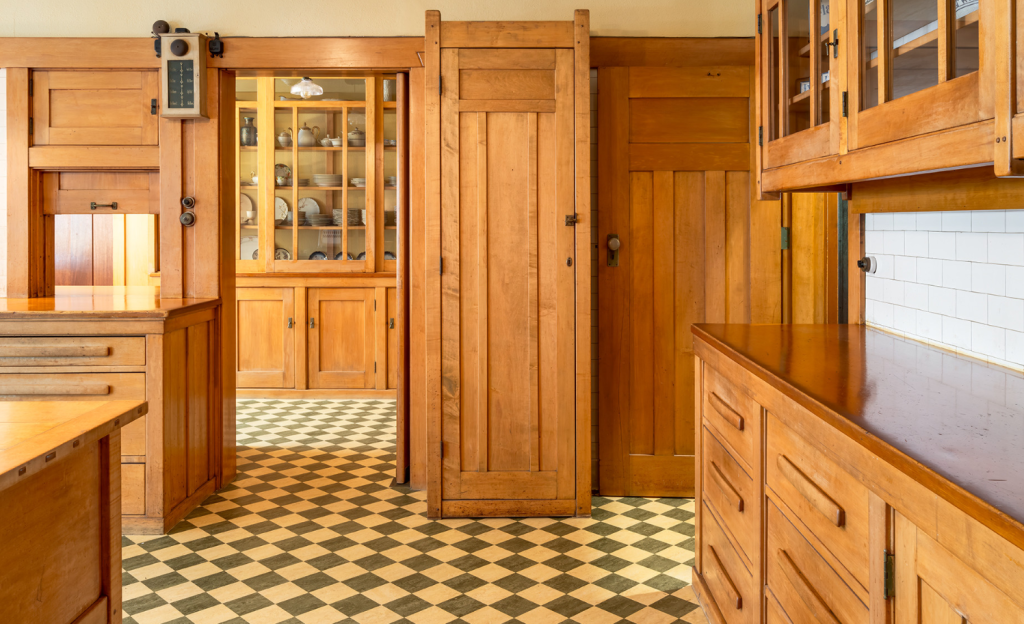 Alternate view of the Gamble House kitchen photographed by Alexander Vertikoff showing the butler's pantry area with glass-fronted Honduras mahogany china cabinet displaying dishware on multiple shelves with geometric panel designs on the lower cabinet doors, Honduras mahogany pocket door at center with vertical board-and-batten panel design, white painted cabinetry at right with geometric panel designs, checkered linoleum flooring in cream and sage green diagonal pattern continuing throughout the space, white subway tile backsplash, vintage wall-mounted telephone and electrical panel visible on the left wall, and warm golden lighting creating functional service spaces that maintain the aesthetic integrity and honest expression of materials found throughout the house while serving the practical needs of early twentieth-century domestic life