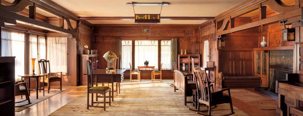 Living room of the Gamble House photographed by Alexander Vertikoff showing the complete spatial composition with Honduras mahogany wall paneling throughout, exposed beam ceiling with dark-stained beams and cream plaster between creating a coffered pattern, massive structural posts with cloud-lift brackets supporting the ceiling beams, hand-carved frieze panels depicting gnarled oak trees with organic forms running along the upper walls, custom pendant light fixture with geometric design and amber glass panels suspended from the ceiling, Greene and Greene library table by Peter Hall Manufacturing Company with ebony pegs and drawers surrounded by chairs with vertical slat backs and upholstered seats, inglenook fireplace area visible at left with built-in bench and green cushion, Tiffany table lamp with leaded glass shade, custom upright piano with Honduras mahogany case at right, Arts and Crafts carpet with floral and geometric patterns, hardwood flooring, and warm golden afternoon light streaming through windows creating an intimate and contemplative living space that exemplifies the ultimate bungalow aesthetic