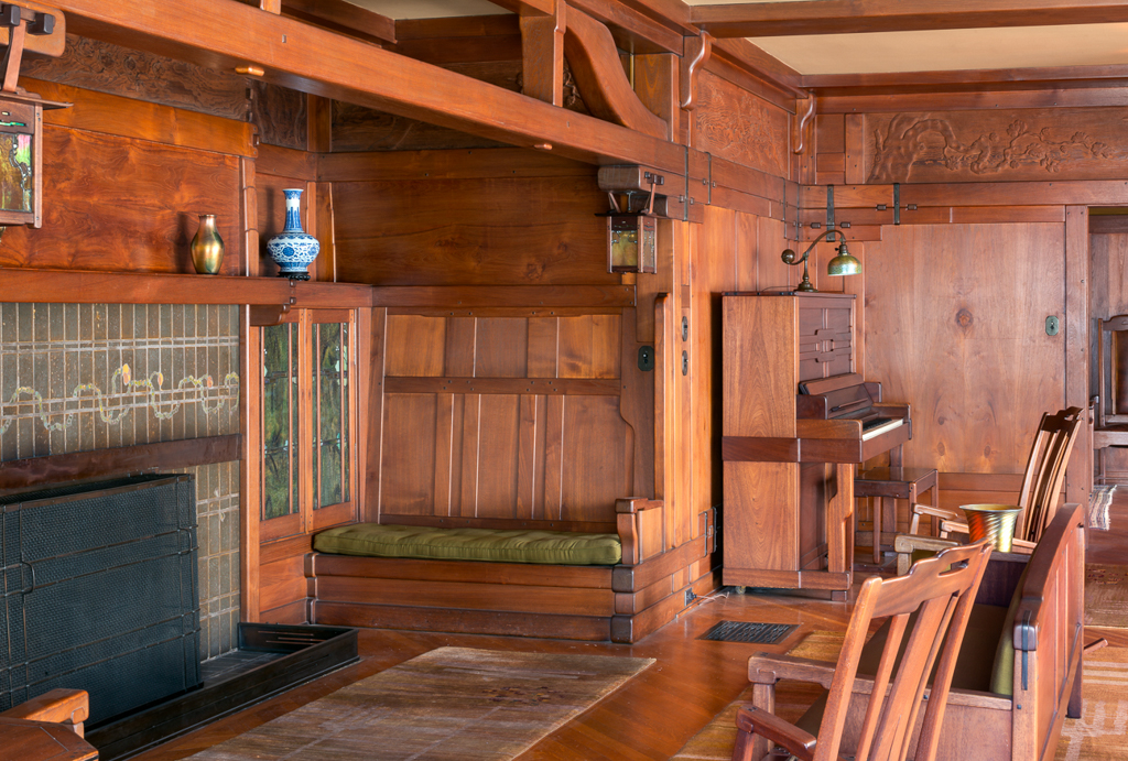 Inglenook area of the Gamble House living room photographed by Alexander Vertikoff showing the built-in fireplace with iridescent green tile surround in matte finish, Honduras mahogany built-in bench with green velvet upholstered cushion positioned beside the fireplace, leaded glass cabinet doors with geometric pattern displaying pottery on shelves, hand-carved frieze panels depicting gnarled oak trees running along the upper walls, massive exposed structural beams and posts with cloud-lift brackets, custom pendant light fixtures with wrought iron hardware suspended from the ceiling, custom upright piano with Honduras mahogany and bird's-eye maple case visible at right, Greene and Greene chairs with vertical slat backs, hardwood flooring, and the warm golden lighting creating an intimate inglenook seating area that invites contemplation beside the hearth