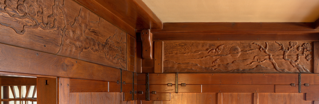 Close-up detail of the hand-carved Honduras mahogany frieze panels in the living room of the Gamble House showing stylized gnarled oak tree with twisted trunk, spreading branches, and organic forms carved in low relief, the frieze running continuously along the upper wall beneath the exposed beam ceiling, custom Honduras mahogany cabinetry below with geometric panel designs and decorative metal strap hinges with hammered finish, small windows with V-shaped muntins providing natural light, and the warm golden-brown patina of aged wood demonstrating the integration of decorative arts with architectural elements characteristic of Greene and Greene's ultimate bungalow designs
