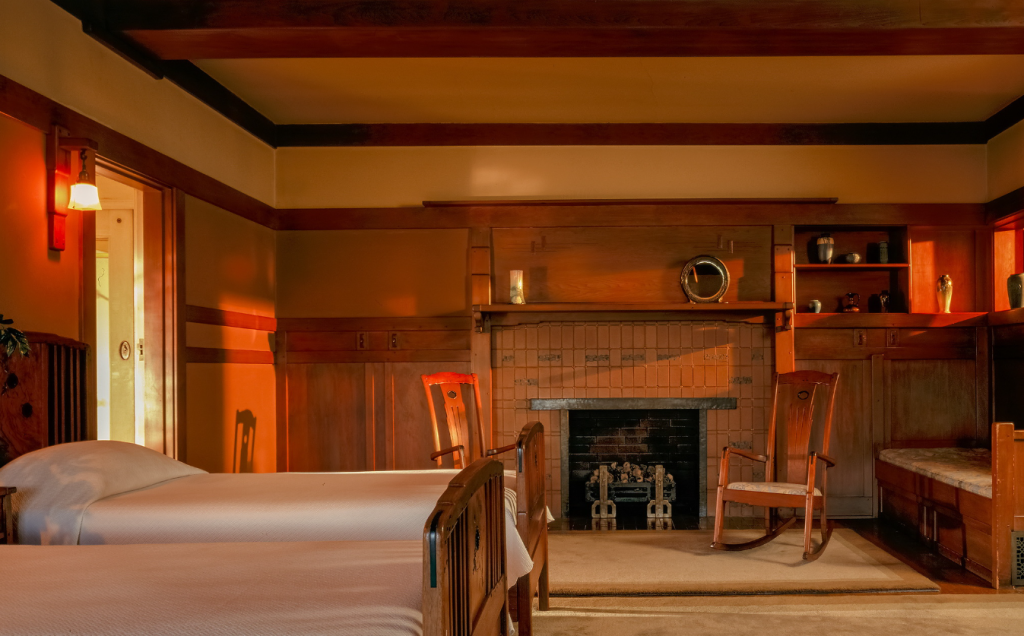 Alternate view of the master bedroom at the Gamble House showing the spatial flow from the bedroom through Honduras mahogany pocket doors to the sleeping porch beyond where windows with V-shaped muntins frame views of mature oak trees in warm sunlight, eight-drawer dresser with tilting mirror and silver inlay at center flanked by two side chairs with vertical slat backs, Honduras mahogany occasional table at left with books and decorative objects, rocking chair at right, custom pendant light fixture with geometric design and amber glass panels suspended from the exposed beam ceiling, warm peach-toned walls with Honduras mahogany wainscoting creating horizontal bands, Arts and Crafts wall sconces with amber glass shades, cream-colored carpet, built-in cabinetry visible at far left, and the warm afternoon light streaming through creating connected indoor-outdoor living spaces characteristic of the California bungalow lifestyle