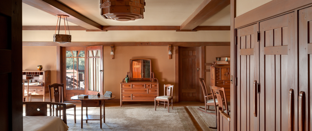 Master bedroom at the Gamble House at sunset showing twin Honduras mahogany beds with vertical slat headboards and footboards dressed in white coverlets positioned on either side of the built-in fireplace, fireplace with cream-colored tile surround in geometric pattern and black tile hearth with decorative andirons, Honduras mahogany mantel with open shelving above displaying pottery and decorative objects, Honduras mahogany rocking chair and side chair positioned in front of the fireplace on cream-colored area rug, built-in cabinetry flanking the fireplace with geometric panel designs and open shelving, warm peach-toned walls with Honduras mahogany wainscoting and trim creating horizontal bands, exposed beam ceiling with dark-stained beams and cream plaster between, Arts and Crafts wall sconces with amber glass shades providing warm ambient lighting, doorway at left leading to adjacent room, and warm golden sunset light creating an intimate and romantic sleeping space