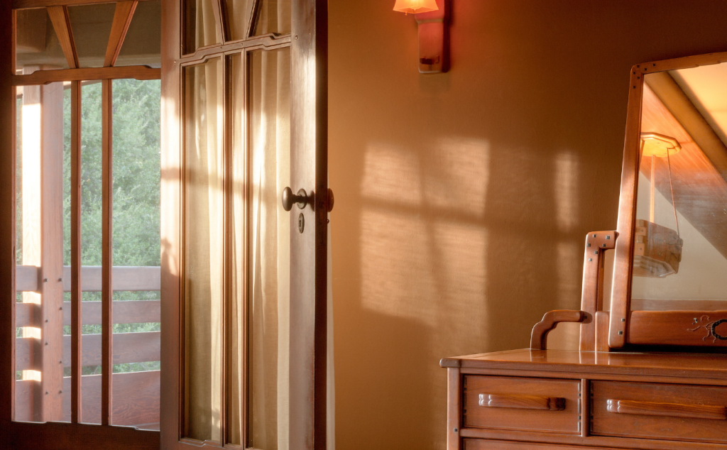 Mahogany door with board-and-batten design opening to sleeping porch with railing and oak tree views, dresser with tilting mirror casting shadows, Arts and Crafts sconce, warm afternoon sunlight creating patterns