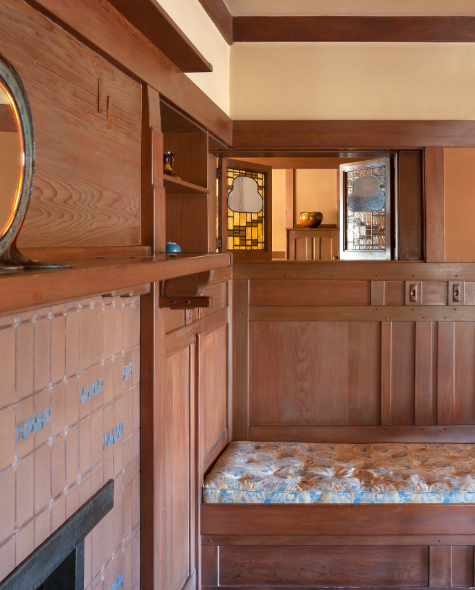 Built-in fireplace with cream tile surround, mahogany bench with floral upholstered cushion, open shelving with pottery, view through doorway to glowing Tiffany stained glass windows in golden-yellow and amber