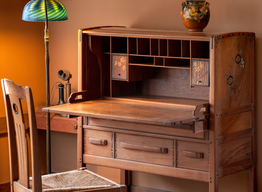 Writing desk by Peter Hall Manufacturing Company in mahogany and bird's-eye maple with drop-front open revealing compartments and drawers, silver and abalone shell inlay on sides, accompanied by chair, Tiffany lamp with blue and green swirled glass dome shade, vintage telephone, pottery vase
