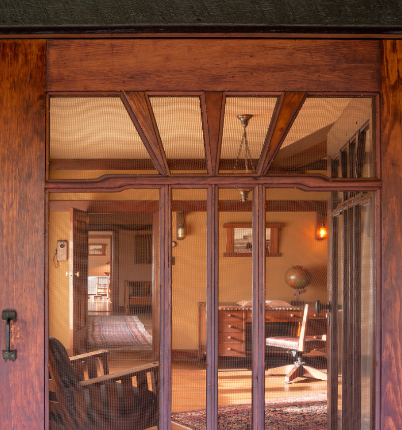 View from boys' bedroom sleeping porch through open door into bedroom, showing Arts and Crafts interior with exposed ceiling beams, pendant lighting, Stickley desk with multiple drawers, and period swivel chair. Woven cane screens in door panels filter light while maintaining transparency between spaces.