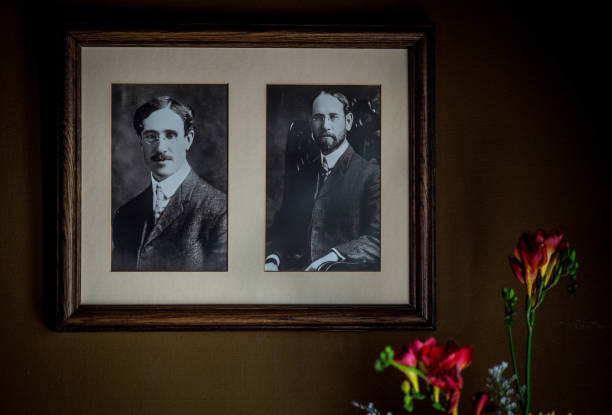 Framed dual portrait of architects Charles Sumner Greene (with mustache) and Henry Mather Greene (with beard) in formal Edwardian attire circa early 1900s, sepia-toned in cream matting within mahogany frame, fresh flowers in foreground
