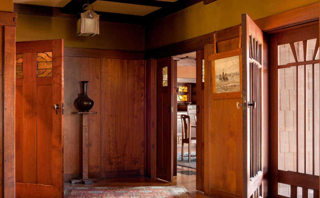 Mahogany paneling with board-and-batten design, pocket door with silver tree inlay, console with bronze vase, door with V-shaped muntins and sheer curtains, framed seascape, view into dining room with glowing Tiffany windows and dining chair, Arts and Crafts pendant light