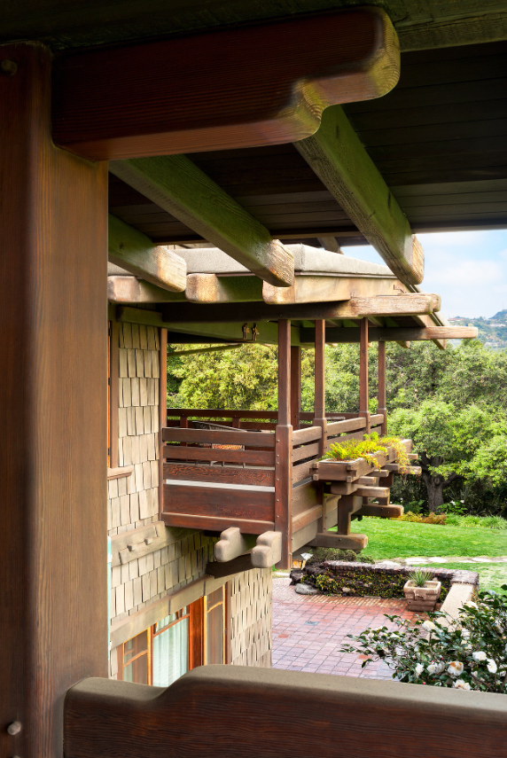 View looking out showing massive exposed beams and posts painted soft green, weathered shingle siding, mahogany railing, windows with V-shaped muntins, views to terraced walls, herringbone brick, plantings, lawn, oak trees, and San Gabriel Mountains