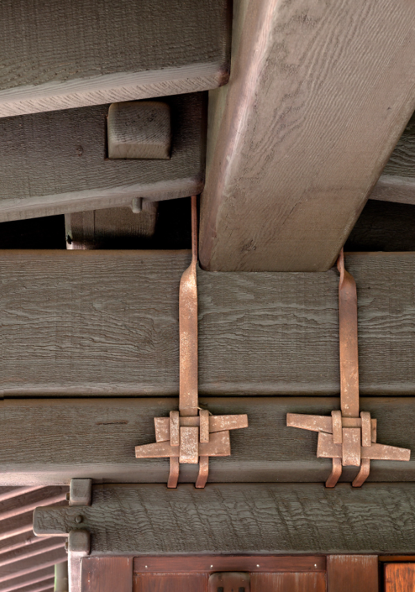 Close-up of structural connection showing massive timber beam painted soft green supported by post, connected with custom copper straps featuring decorative geometric plates with square pegs in Arts and Crafts pattern