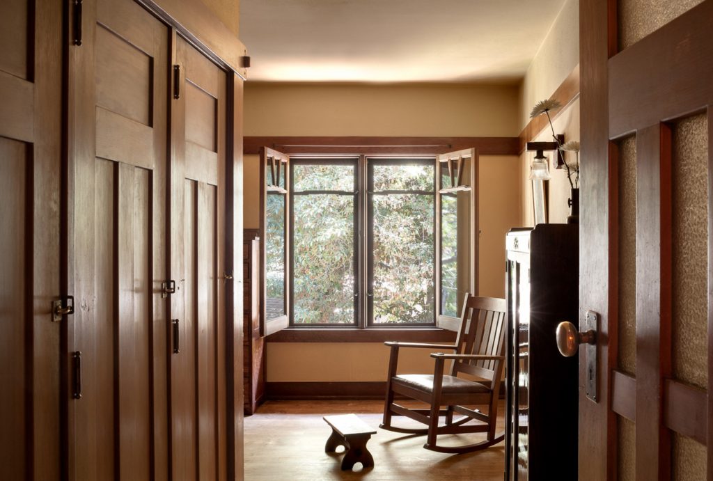 Staff bedroom at the Gamble House showing built-in window seat with Honduras mahogany frame and casement windows opening outward to views of mature oak trees, Honduras mahogany rocking chair with upholstered seat positioned on the window seat, wooden footstool, Honduras mahogany pocket doors at left, warm peach walls with mahogany trim creating horizontal bands, Arts and Crafts wall sconces, hardwood flooring.