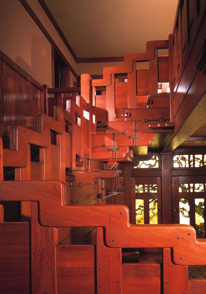 Dramatic view looking up the grand staircase at the Gamble House showing the sculptural cloud-lift balustrade with stepped geometric forms in Honduras mahogany with square ebony pegs creating a rhythmic pattern, Arts and Crafts carpet runner ascending the stairs, art glass windows with stylized oak tree branches in golden-yellow and amber glass glowing at the landing, Honduras mahogany wall paneling, exposed beam ceiling.