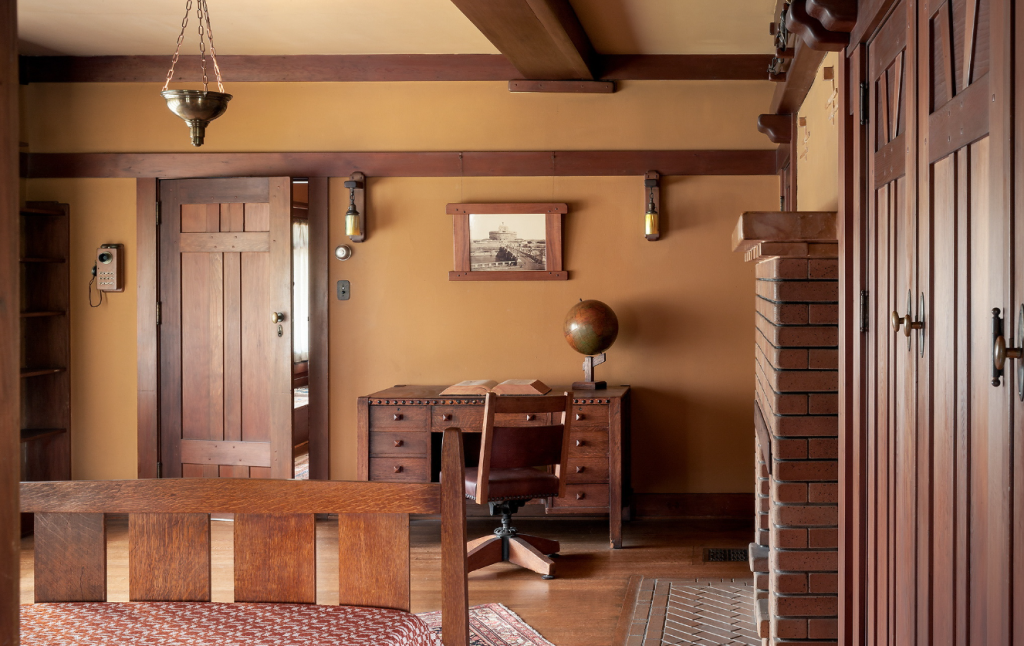 Upstairs bedroom at the Gamble House photographed by Alexander Vertikoff showing Honduras mahogany desk with leather office chair, vintage globe on stand, framed historic photograph with mahogany frame, Arts and Crafts wall sconces with amber glass shades, pocket door with geometric panels leading to adjacent room, brick staircase visible at right, exposed beam ceiling, hardwood flooring, Arts and Crafts carpet.