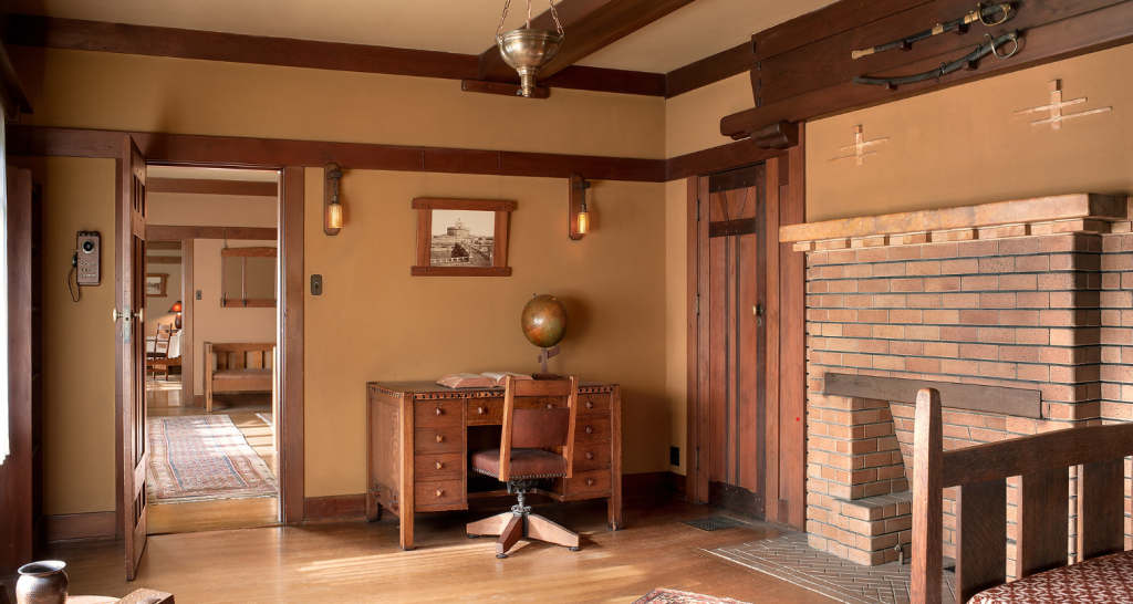 Alternate view of upstairs bedroom at the Gamble House photographed by Alexander Vertikoff showing the dramatic brick fireplace with arched opening constructed in variegated clinker brick with darker headers creating decorative pattern, Honduras mahogany desk with leather office chair and vintage globe, framed historic photograph, Arts and Crafts wall sconces, view through doorway to hallway with bed visible, exposed beam ceiling, custom pendant light fixture.