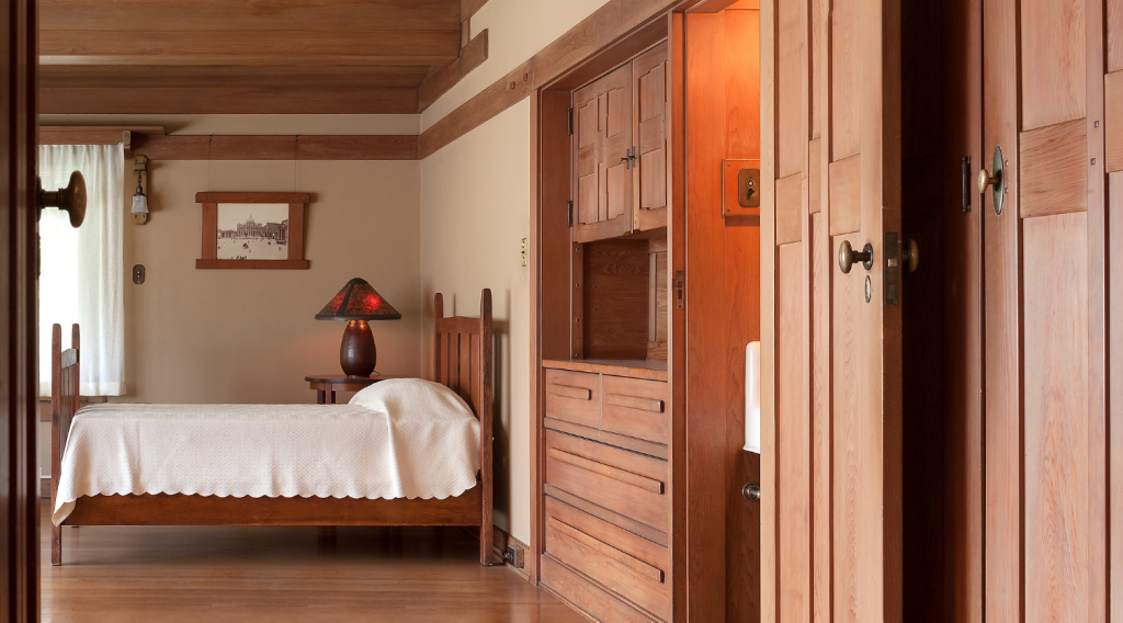 Upstairs bedroom number 2 at the Gamble House showing Honduras mahogany bed with vertical slat headboard and white coverlet, Arts and Crafts table lamp with pyramidal shade in red and amber glass on wooden base positioned on nightstand, framed historic photograph with mahogany frame on wall, Honduras mahogany built-in dresser with geometric panel designs and mirror at center, pocket doors with geometric panels, warm cream walls with mahogany trim, exposed beam ceiling, hardwood flooring.
