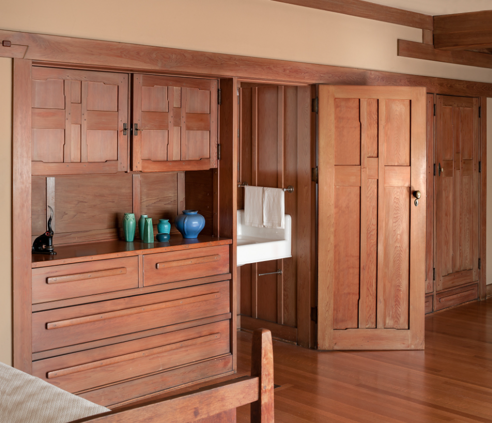 View of upstairs bedroom at the Gamble House showing Honduras mahogany built-in dresser with geometric panel designs displaying turquoise pottery vases, view through pocket door to bathroom with white towel visible, custom pocket doors with geometric panels leading to hallway, warm peach walls with mahogany trim, exposed beam ceiling.