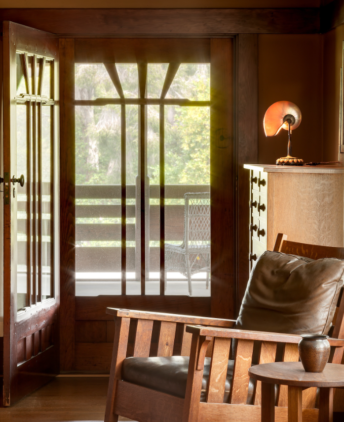 View from upstairs bedroom at the Gamble House photographed by Alexander Vertikoff showing Honduras mahogany French doors with V-shaped muntins opening to the sleeping porch where wicker furniture and railing are visible in warm afternoon sunlight, Honduras mahogany Morris chair with vertical slats and black leather cushions at right, occasional table, nautilus shell table lamp with amber glass shade, warm golden lighting.