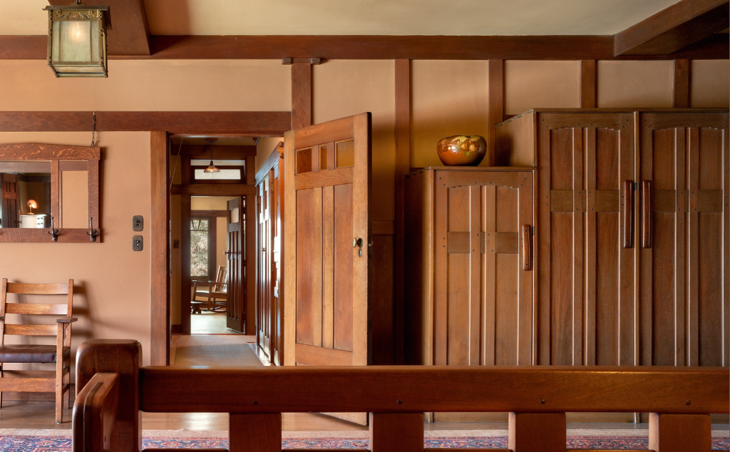Upstairs hall photographed by Alexander Vertikoff showing cloud-lift balustrade with stepped geometric forms and square ebony pegs overlooking the entrance hall below, built-in Honduras mahogany linen closets with geometric panel designs topped with iridescent pottery bowl, Honduras mahogany pocket door opened to reveal view through multiple rooms, Arts and Crafts lantern suspended from ceiling, warm peach walls, exposed beam ceiling.