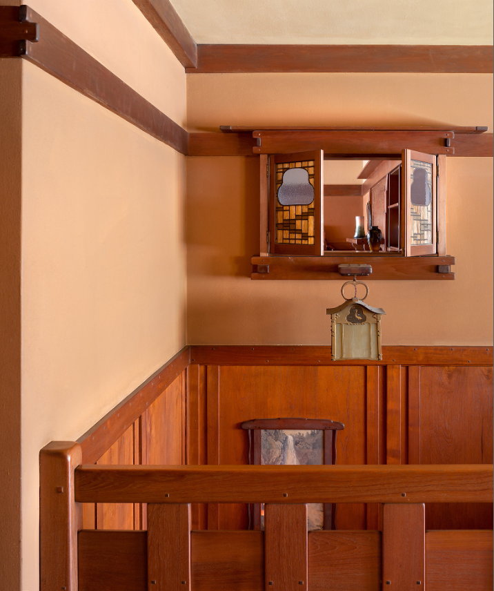 View of upstairs hall showing cloud-lift balustrade with stepped geometric forms and square ebony pegs, built-in ventilation window above with leaded glass featuring cloud-lift design in amber and sage green, Arts and Crafts lantern with amber glass suspended from ceiling, warm peach walls with Honduras mahogany trim, exposed beam ceiling.