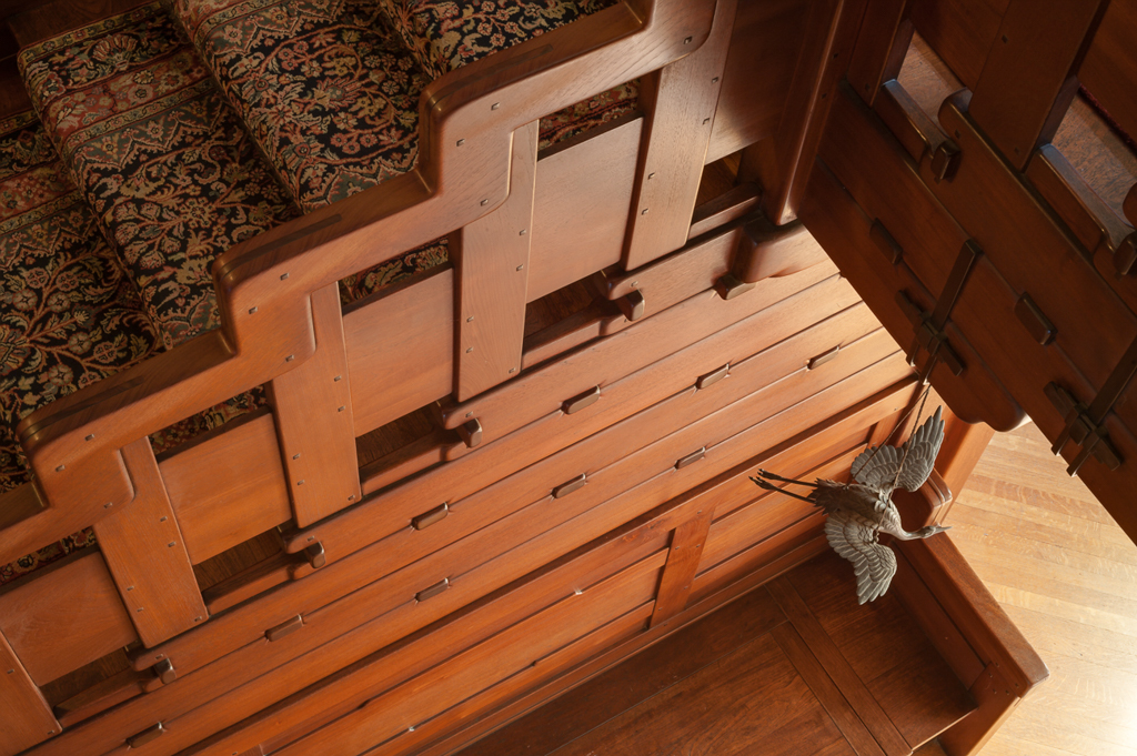 View looking down from upstairs hall balcony showing the cloud-lift balustrade with stepped geometric forms in Honduras mahogany with square ebony pegs and custom copper straps with decorative plates, Arts and Crafts carpet with floral and geometric patterns below, bronze bird sculpture perched on the railing, demonstrating Greene and Greene's integration of decorative elements with structural design.