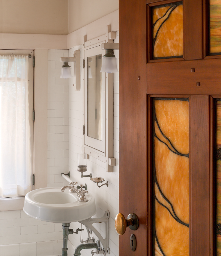 View through Honduras mahogany door with leaded art glass panel depicting stylized Japanese tsuba shapes in golden-amber glass into white-tiled bathroom showing wall-mounted porcelain sink with nickel-plated faucets, medicine cabinet with mirror flanked by Arts and Crafts sconces, window with sheer curtains, white subway tile walls, custom bronze door handle.