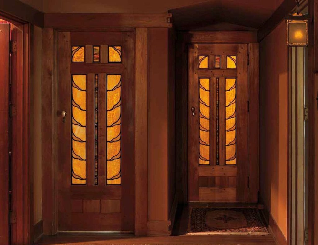 Two Honduras mahogany doors in upstairs hall glowing with warm backlighting, each featuring leaded art glass panels with stylized Japanese tsuba shapes in golden-amber glass with darker leading creating silhouettes, geometric panels in upper transom sections, warm peach walls, Arts and Crafts lantern with amber glass at right, Arts and Crafts carpet.