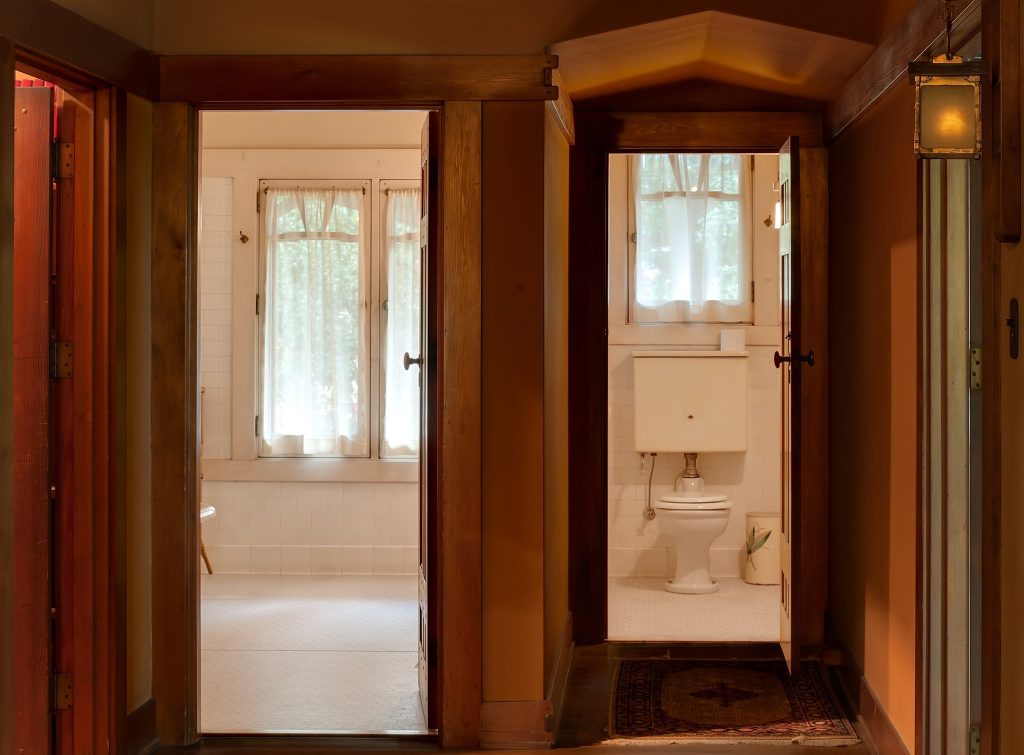 Two Honduras mahogany doors opened to reveal white-tiled bathroom at left with window and sheer curtains, and alternate bathroom at right with white porcelain toilet visible, white walls with hand-rubbed trim, exposed beam ceiling with tongue-and-groove wood between, Arts and Crafts lantern, Arts and Crafts carpet runner.