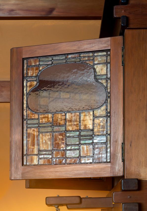 Custom Honduras mahogany ventilation window in upstairs hall with leaded glass panel featuring cloud-lift organic form in purple-gray glass surrounded by geometric border of golden-amber and sage green glass in Arts and Crafts pattern, mounted in mahogany frame with custom hinges, warm golden-yellow walls visible.