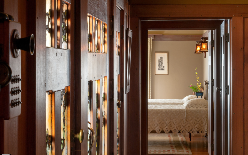 View through open Honduras mahogany doorway into guest bedroom with white crocheted bedspread, framed artwork, and wall-mounted sconce with amber glass shade. Foreground shows illuminated art glass panels in adjacent entrance hall front door with decorative hardware, demonstrating layered spatial composition and integrated lighting design throughout the house.