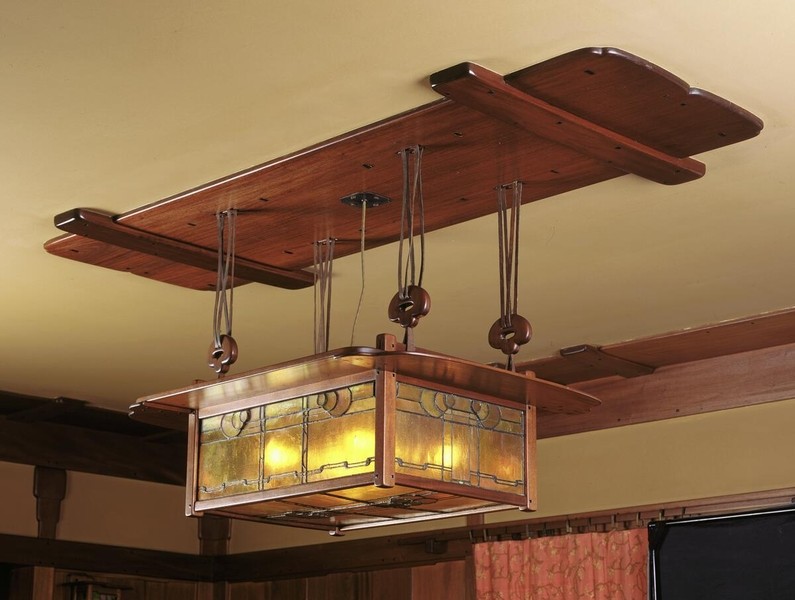 Substantial dining room chandelier with sculptural wooden canopy featuring curved wing-like extensions. The rectangular fixture displays amber and green art glass panels with stylized plant motifs in copper-toned metal frame, suspended by adjustable pulley system with decorative knots and hardware.