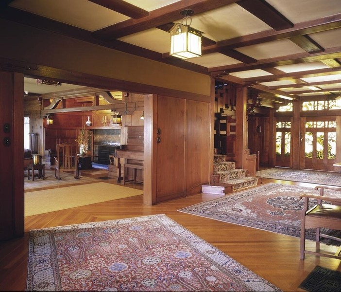 Wide-angle view of entrance hall interior showcasing Arts and Crafts architecture with exposed beam ceiling, polished wood floors, and Oriental rugs. Multiple lantern-style pendant fixtures illuminate the space, with views through doorways to adjacent living areas featuring built-in furniture and craftsman woodwork details.