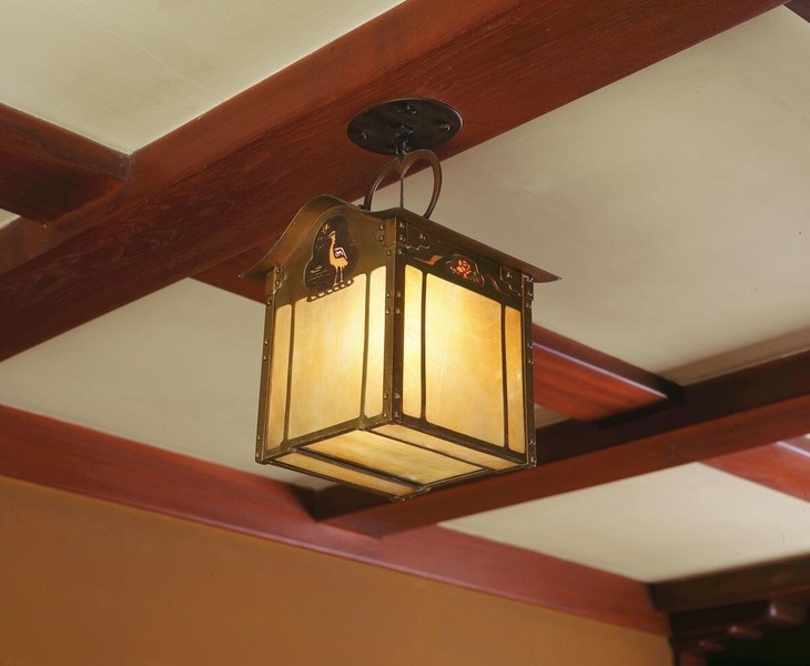 Lantern-style pendant fixture with square form, featuring amber glass panels within dark metal frame with decorative cutout details. Suspended by curved metal loop from circular ceiling mount, positioned beneath exposed wooden beams painted in rich red-brown tones typical of Craftsman interiors.