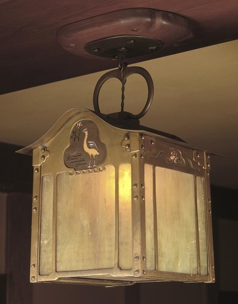 Arts and Crafts lantern-style ceiling fixture with gold-plated metal frame and translucent amber glass panels. Features decorative bird motif at top, riveted construction details, and curved loop suspension hardware mounted to dark wood ceiling, exemplifying Greene and Greene's attention to handcrafted detail.